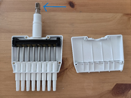 A disassembled white Pipetman laboratory pipette with an arrow pointing to the top metal section, all placed on a wooden surface.