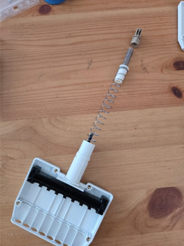 A white plastic Pipetman device with a long metal spring and cylindrical components rests on a wooden surface, partially assembled with several compartments visible.