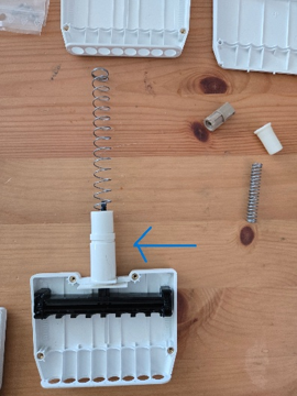 A disassembled Pipetman with visible springs and components lies on a wooden surface; a blue arrow points to a central white cylindrical part.