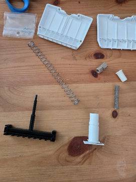 Disassembled plastic and metal components, including springs, screws, casing parts, and Pipetman sections, laid out on a wooden surface.