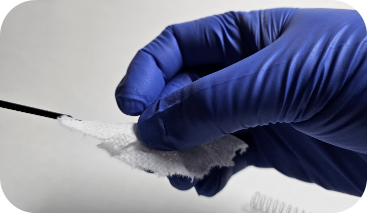 A gloved hand holds a piece of gauze, cleaning or wiping a Pipetman piston against a plain background.