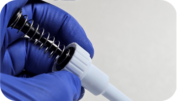 A gloved hand holds a Pipetman piston assembly, aligning a metal spring with a tip holder against a plain background.