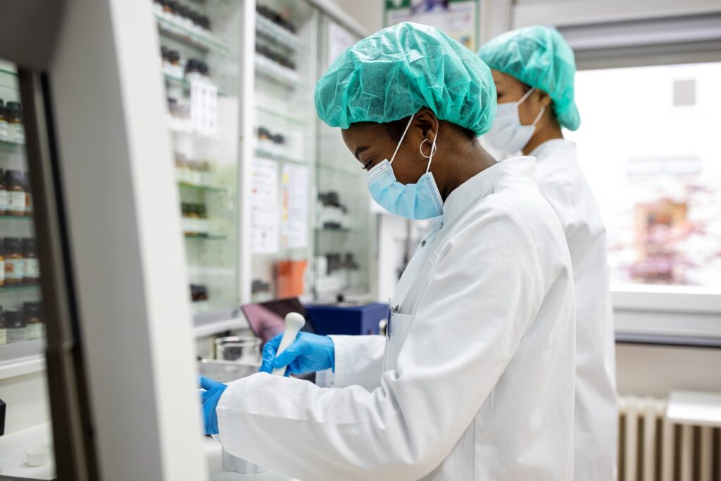 Two scientists wearing protective gear work with lab equipment and samples in a laboratory setting, ensuring compliance with pipette calibration regulations.